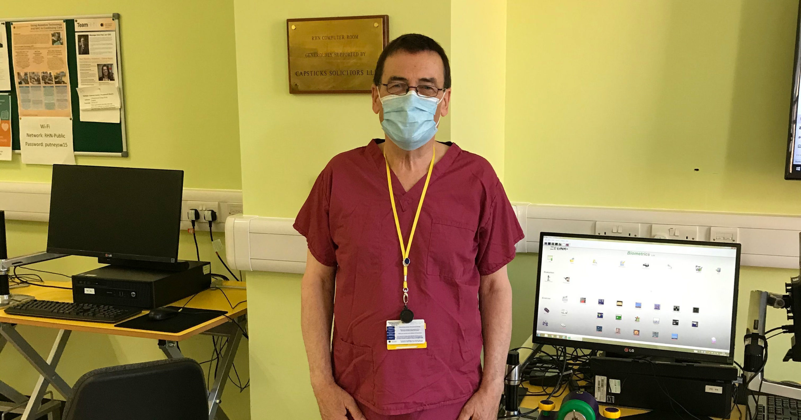 man wearing a mask and lanyard wearing burgundy scrubs standing in front of a computer