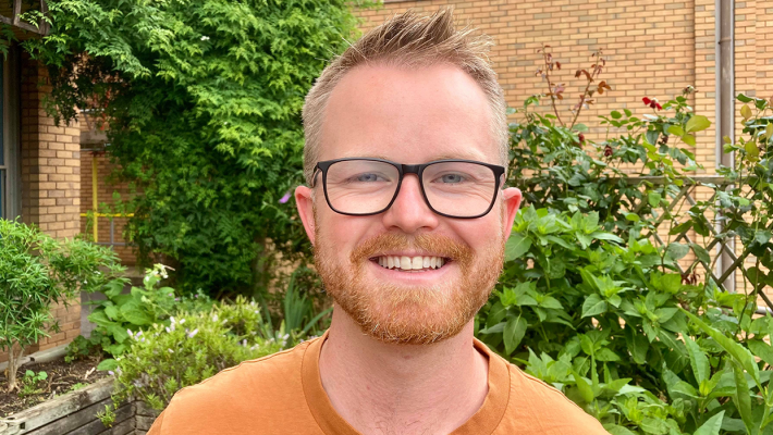 Smiling millennial man with red beard and hair, wearing glass and orange tee, standing in front of green bushes and a brick wall 